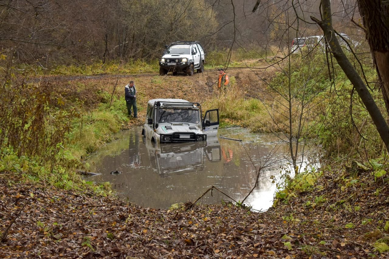 SUV hoisted in muddy water on a forest trail, surrounded by people and lush greenery.