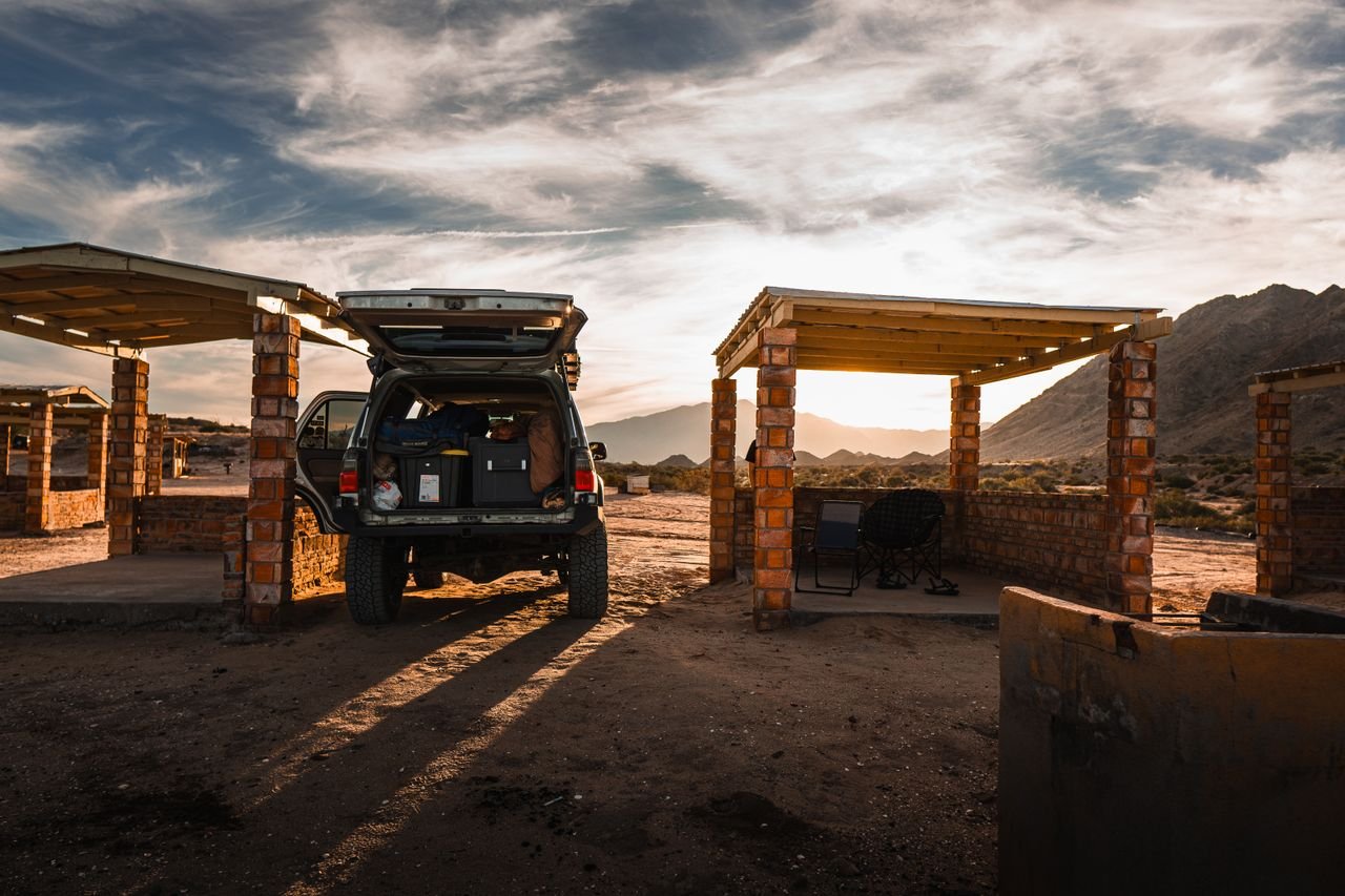 An off-road vehicle parked near rustic gazebos in a mountainous rural setting during sunset.