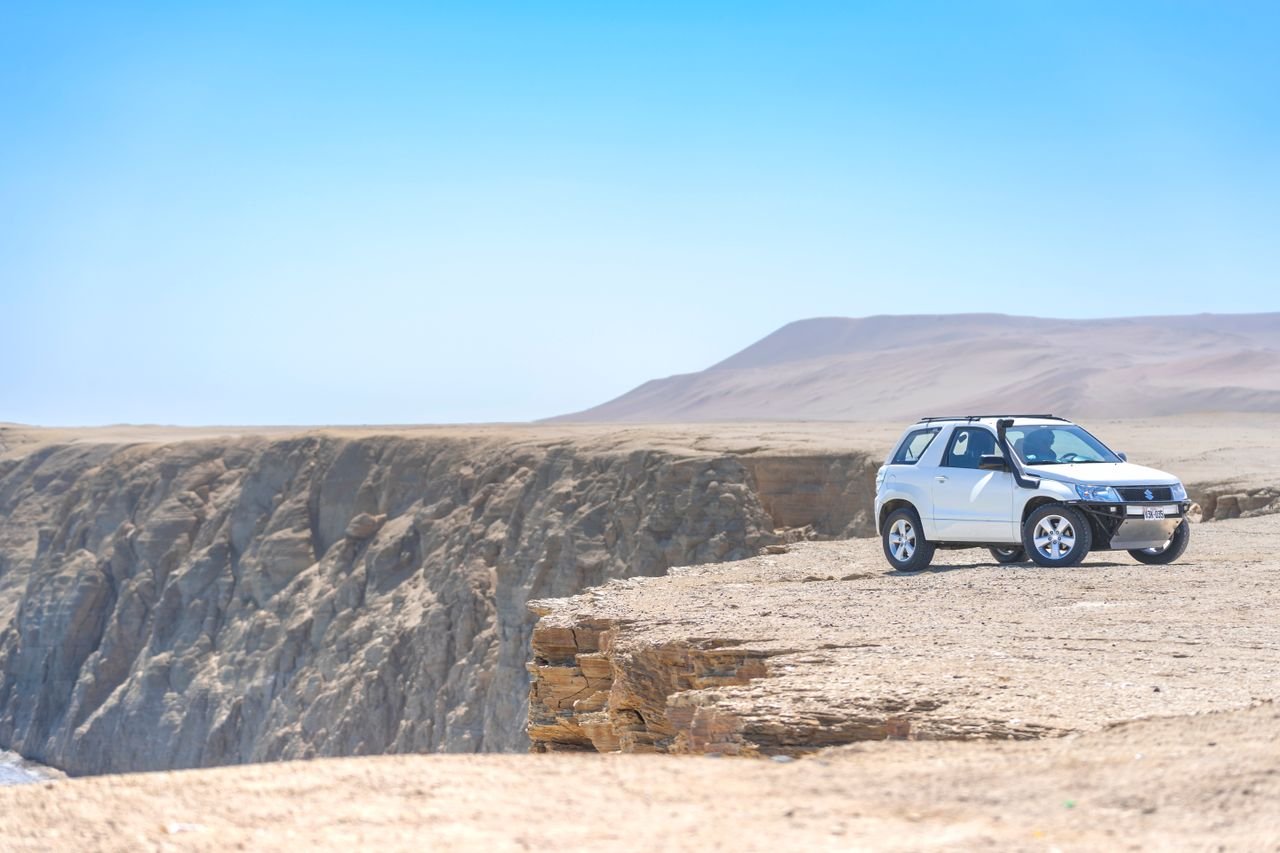 White SUV parked on a cliff edge in a vast desert landscape under a clear blue sky.