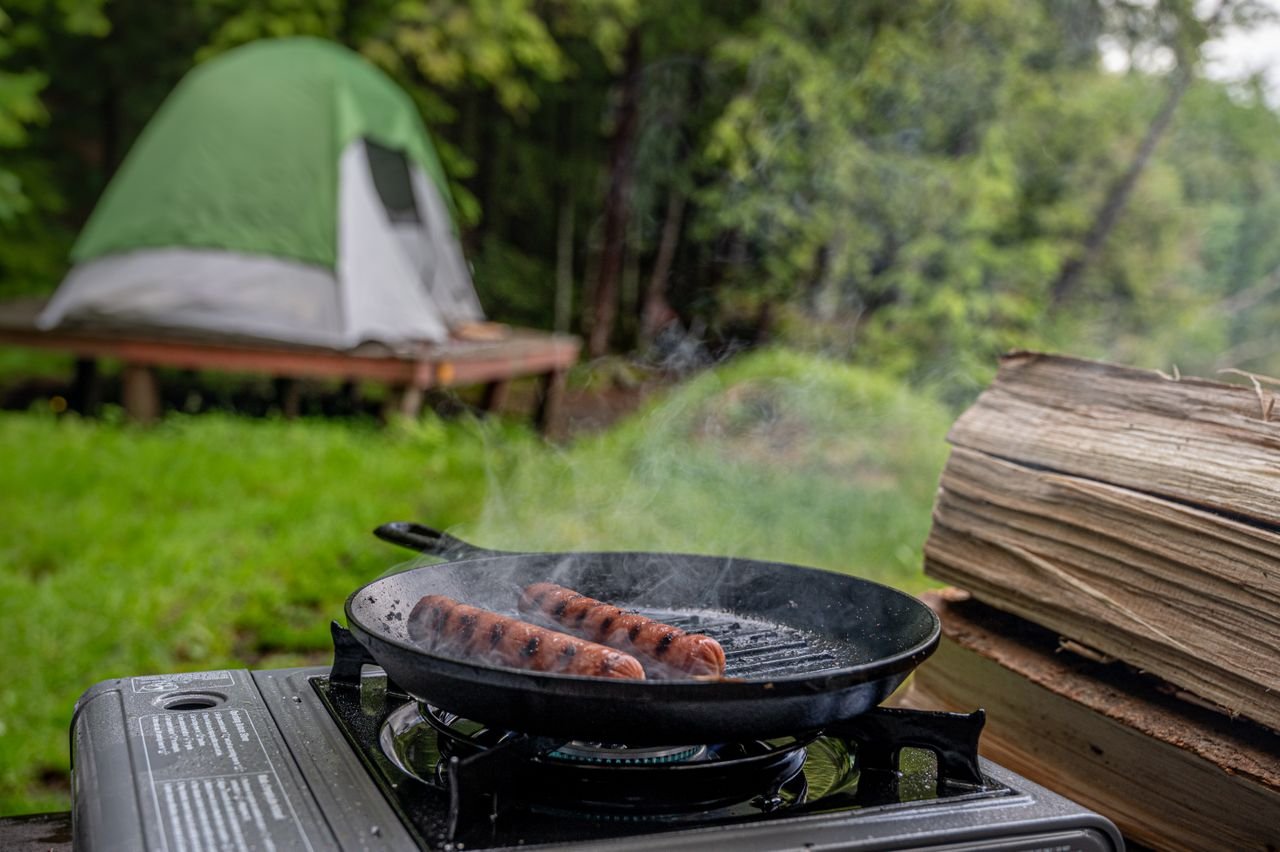 Sausages grilling on a stovetop at a campsite with a tent in the background.