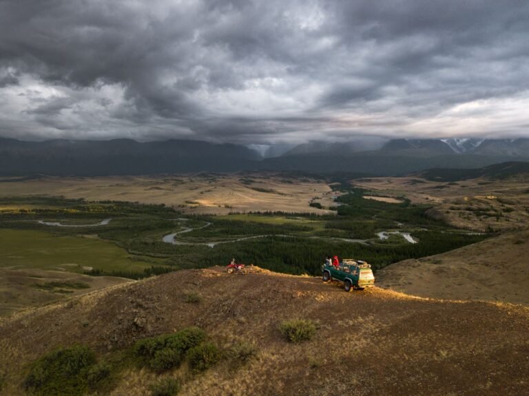 Breathtaking aerial view of unrecognizable travelers and old green car parked on hill slope among green fields and desert valley under dramatic sky