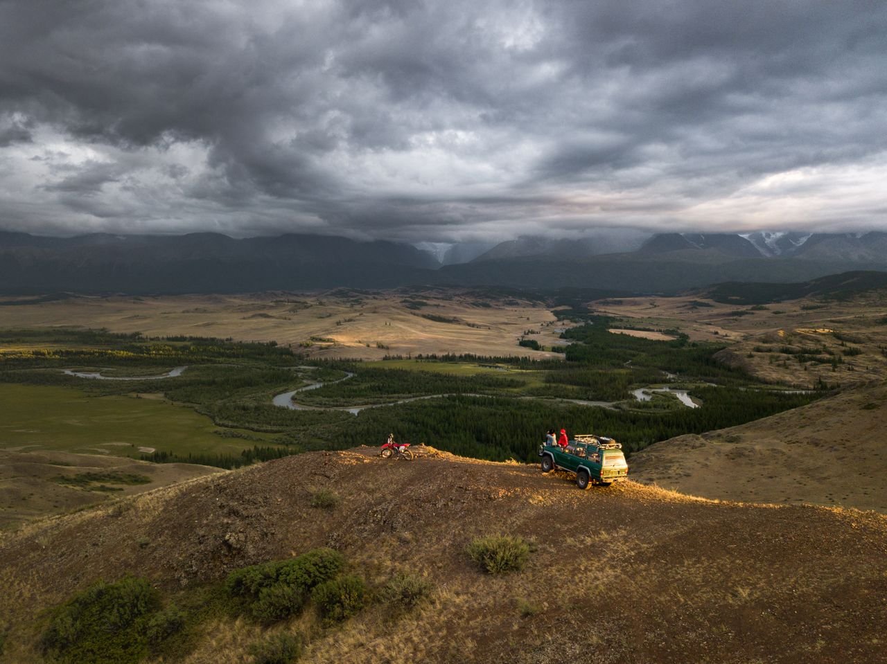 Breathtaking aerial view of unrecognizable travelers and old green car parked on hill slope among green fields and desert valley under dramatic sky