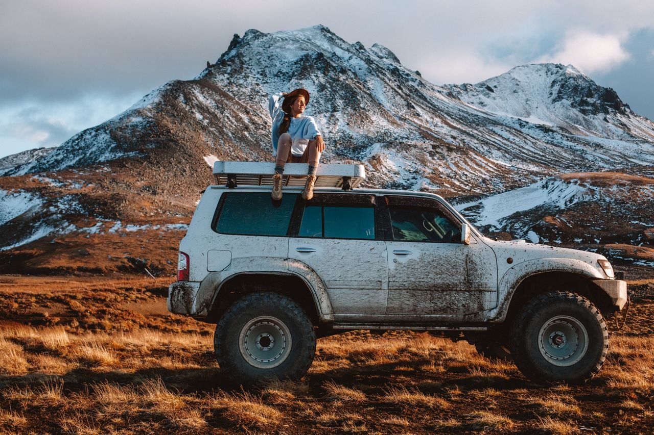 Full body woman in casual clothes and hat sitting on offroader rooftop against snowy mountain range in sunny cloudy day