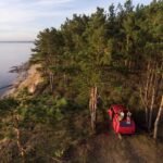 Drone captures a red van parked near a forest cliff overlooking a serene coastline.