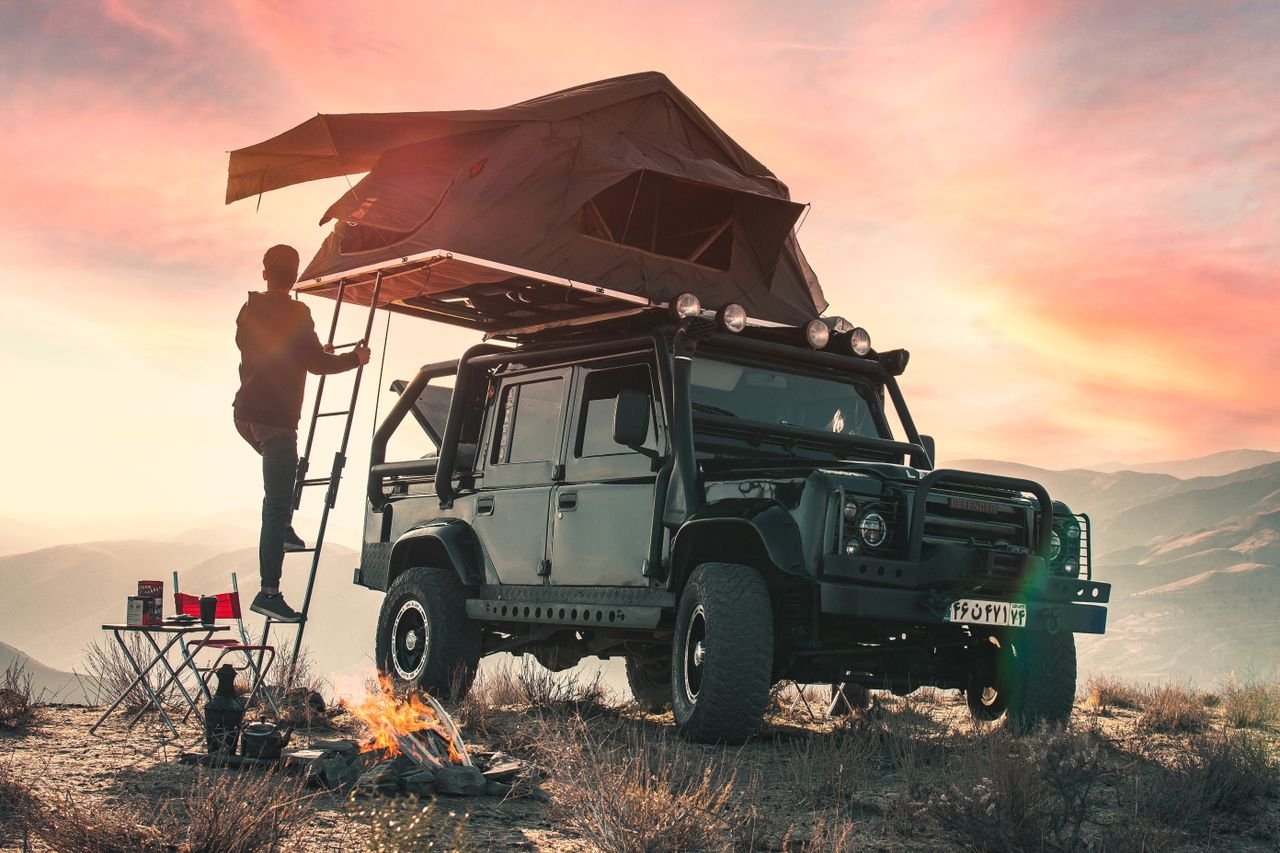 Camping tent placed on roof of SUV car parked on grassy shore near peaceful lake reflecting lush green forest on sunny day