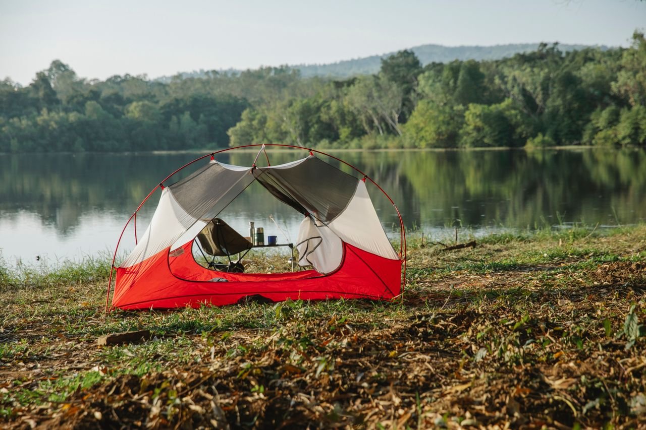 Scenery view of tent on grass coast against lake reflecting green trees under light sky on sunny day