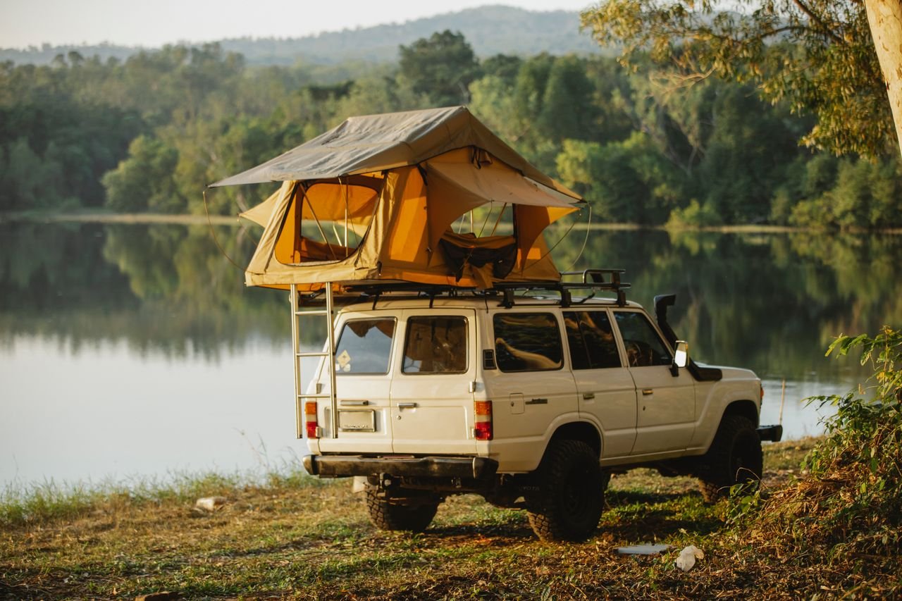 Camping tent placed on roof of SUV car parked on grassy shore near peaceful lake reflecting lush green forest on sunny day